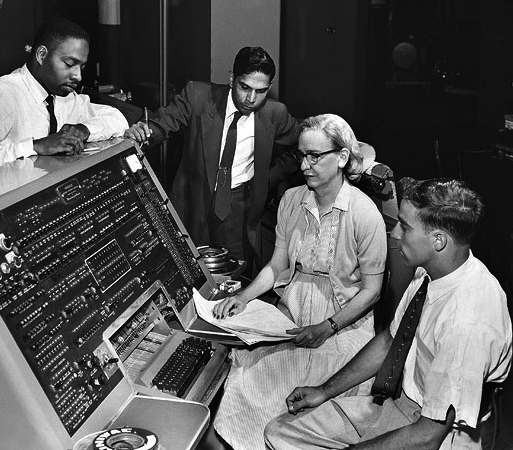 Grace Hopper seated at the UNIVAC console with a manual open on her lap, three colleagues standing around her.