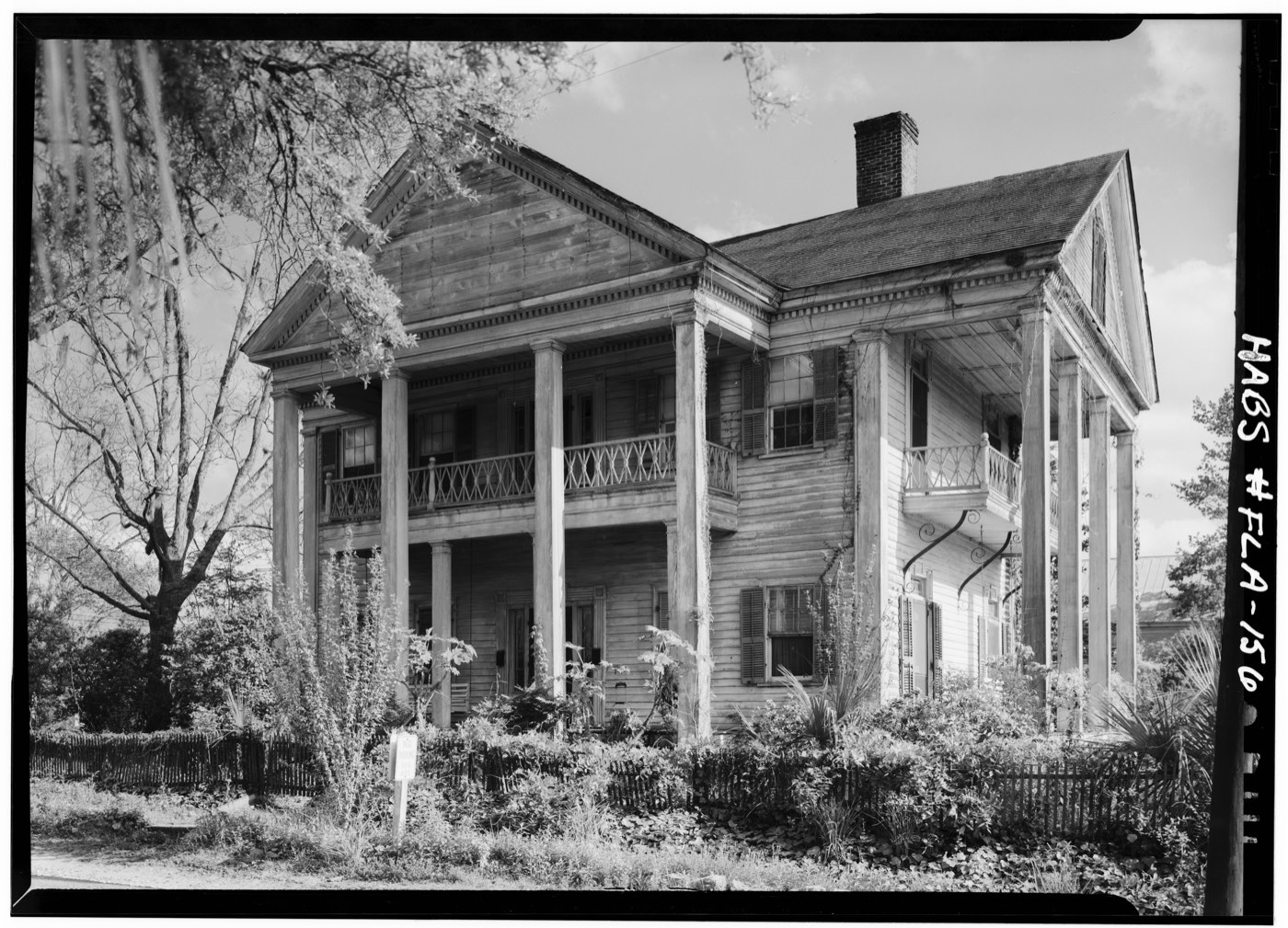 Black and white photograph of the same house, weathered and overgrown, with vines climbing the columns and shrubs across the front.