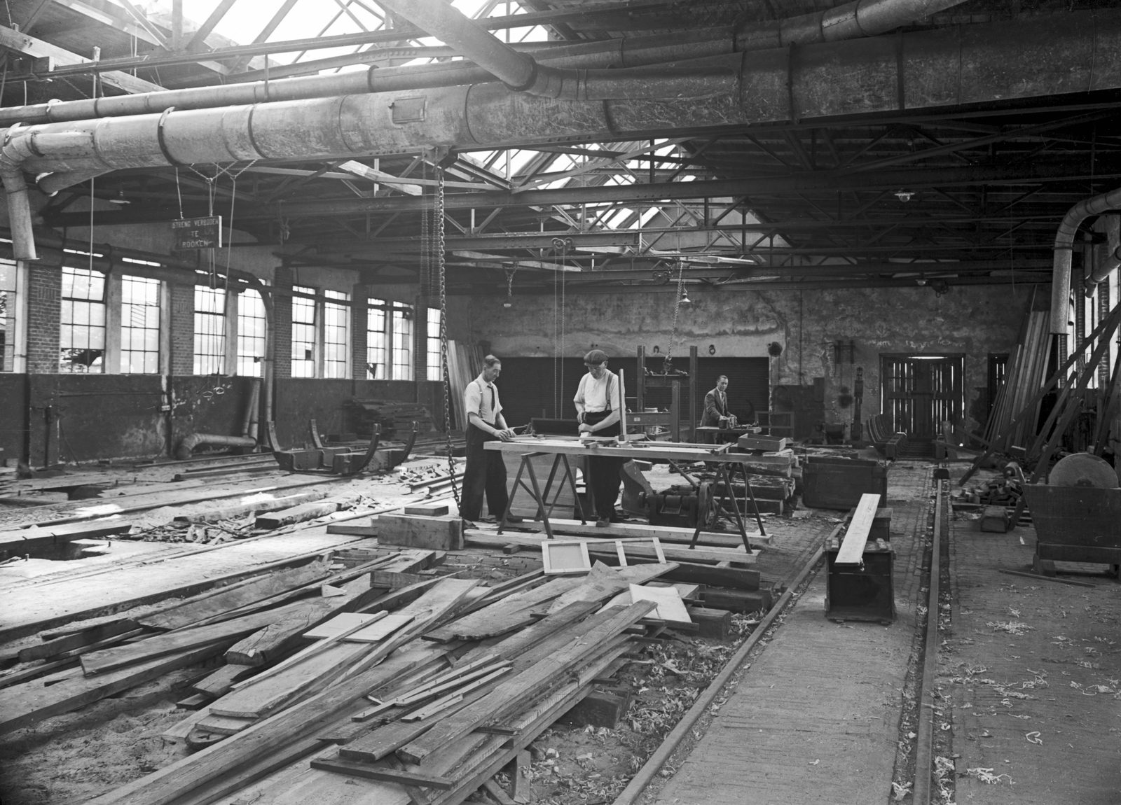 The interior of a large railway woodworking shop, with stacks of sawn timber in the foreground, workbenches down both sides and men working in the distance under a high roof.